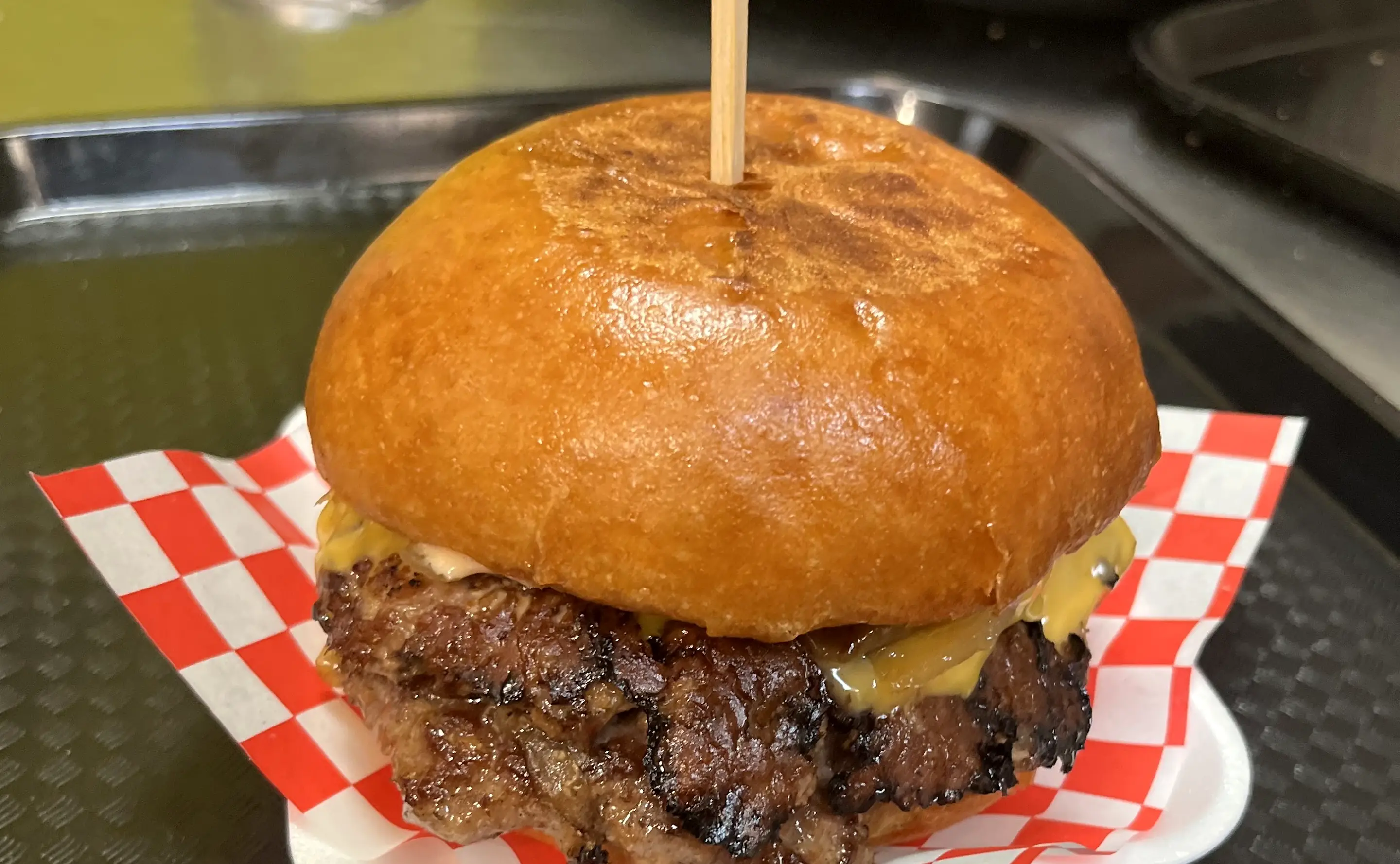 Smash burger with crispy-edged beef patty, melted cheese and grilled onions on a toasted brioche bun with a wooden skewer, on red and white checkered paper