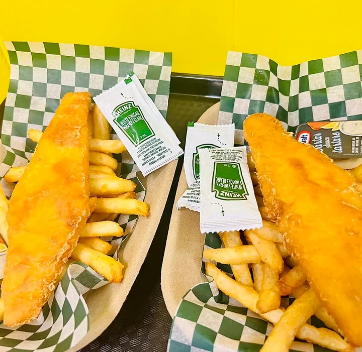 Two pieces of battered fish and chips in paper boats with vinegar and tartar sauce packets